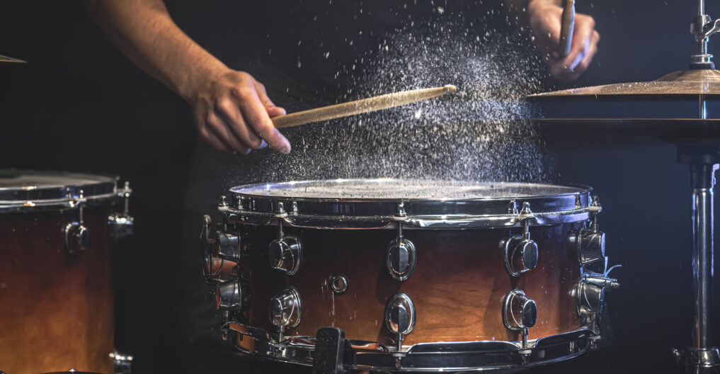 A male drummer plays snare drum with drumsticks in a dark room.